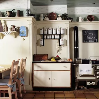 Eine gemütliche Küche mit einem Holztisch und Stühlen sowie einer nostalgischen Kochstelle. An der Wand sind Regale mit Gewürzen und Küchenutensilien dekorativ angeordnet.