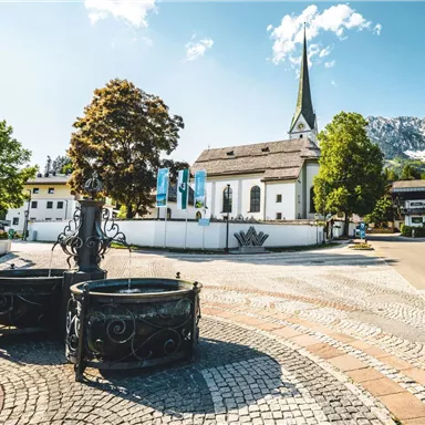 Ein malerischer Dorfplatz mit einer Kirche im Hintergrund und einem klaren Himmel. Umgeben von Bäumen und historischen Brunnen, strahlt die Szene Ruhe und Idylle aus.