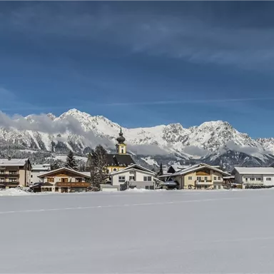 Eine schneebedeckte Landschaft mit einem kleinen Dorf und Bergen im Hintergrund. Der Himmel ist klar und blau.
