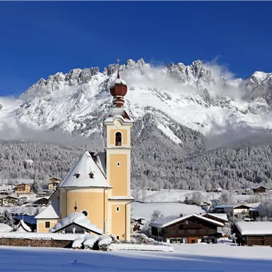Eine malerische Berglandschaft mit schneebedeckten Bergen und strahlend blauem Himmel. Im Vordergrund steht eine charmante Kirche mit einem charakteristischen Turm.