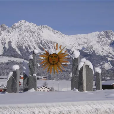 Eine beeindruckende winterliche Landschaft mit schneebedeckten Bergen. Im Vordergrund steht eine Skulptur in Form einer Sonne zwischen steinernen Säulen.
