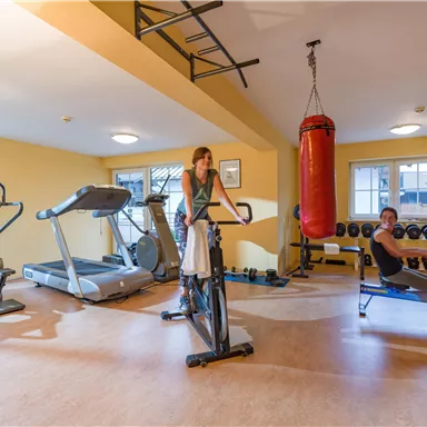 Ein moderner Fitnessraum mit verschiedenen Geräten. Eine Frau trainiert auf einem Ergometer und ein Mann nutzt eine Ruderbank.