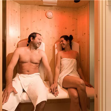 Ein Paar sitzt entspannt in einer Sauna. Sie lächeln sich an und genießen die warmen Holzverkleidungen.