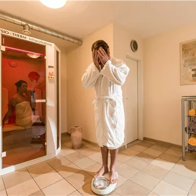 Ein entspannender Wellnessbereich mit einem Mann in einem Bademantel, der auf einer Waage steht. Im Hintergrund ist eine Infrarotkabine sichtbar.