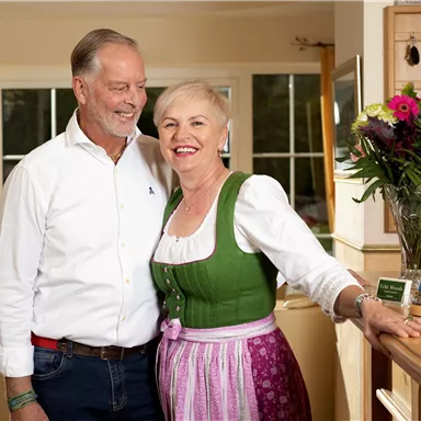 Ein lächelndes Paar steht in einer warmen, einladenden Umgebung. Die Frau trägt ein traditionelles bayerisches Outfit und es gibt Blumen auf der Theke im Hintergrund.