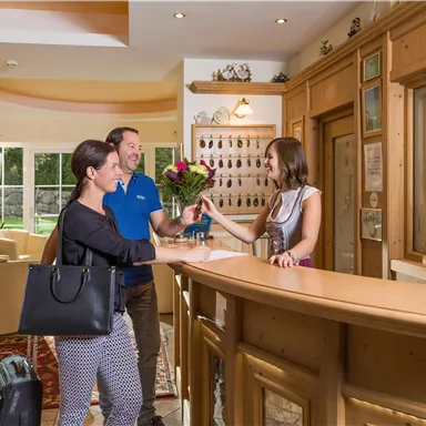 A friendly reception team welcomes a couple at the reception. In the background, cozy seating areas and a bright, inviting atmosphere can be seen.