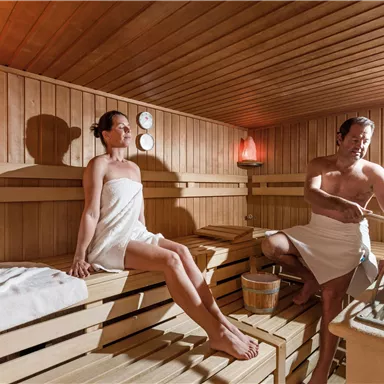 Eine Sauna mit zwei Personen, die entspannen. Die Wände sind aus Holz, und es gibt sanftes Licht von Salzlampen.