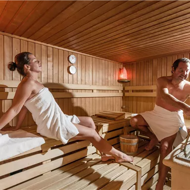 Ein modernes Saunainnenraum mit Holzverkleidung. Zwei Personen entspannen sich in Handtüchern, während sie die Wärme genießen.