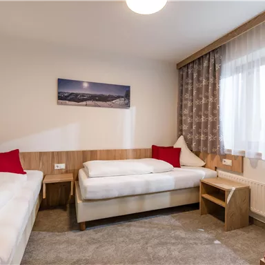 Ein gemütliches Zimmer mit zwei Einzelbetten und einem kleinen Nachttisch. Große Fenster sorgen für viel Tageslicht und eine alpine Landschaft schmückt die Wand.
