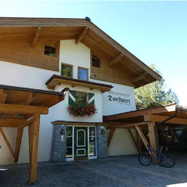 Ein gemütliches Apartmenthaus mit Holzverkleidung und blühenden Pflanzen. Im Vordergrund stehen ein Auto und ein Fahrrad.