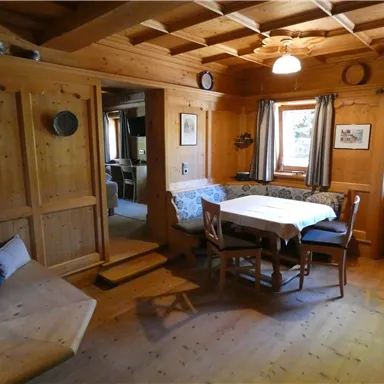 Ein gemütlicher Raum mit Holzwänden und einem großen Tisch. Im Hintergrund sind Fenster mit Vorhängen und ein weicher Sitzbereich zu sehen.