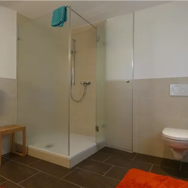 Ein modernes Badezimmer mit einer gläsernen Dusche und einem kleinen Holzstuhl. Die Wände sind in neutralen Farben gehalten, und orangefarbene Handtücher liegen auf dem Boden.