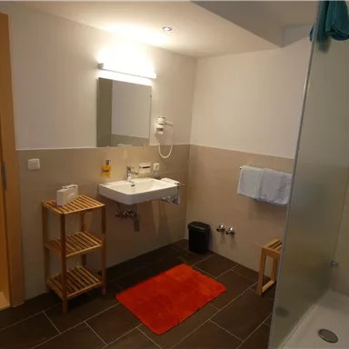 Ein modernes Badezimmer mit hellen Fliesen und einem großen Spiegel. Es gibt eine Dusche, ein Waschbecken und einige Holzmöbel zur Aufbewahrung.