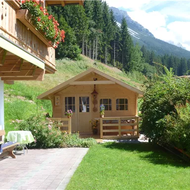 Ein gemütliches Holzhaus in einer grünen Umgebung mit Bergen im Hintergrund. Der sonnige Garten lädt zum Entspannen ein.