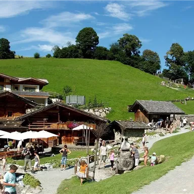 A picturesque landscape with traditional wooden houses and a green meadow. Many people are enjoying the sun and the surroundings.