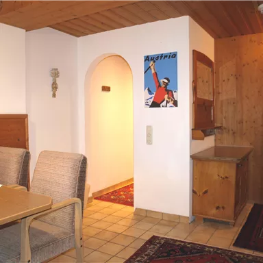 Ein gemütlicher Innenraum mit einem Tisch und Stühlen. Im Hintergrund sieht man eine gemütliche Nische und eine Holztruhe.