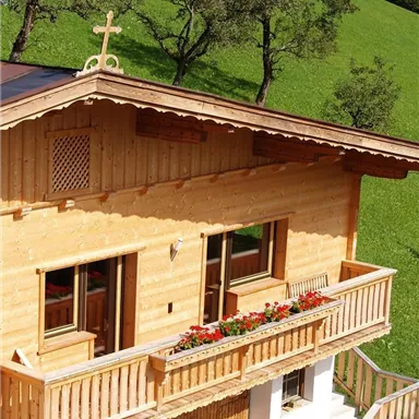 A beautiful wooden house with a balcony full of flowers. In the background, green meadows and trees can be seen.