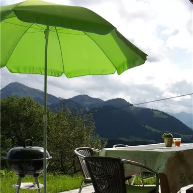 A cozy outdoor area with a green sunshade and a grill. In the background, gentle hills and a cloudy sky can be seen.
