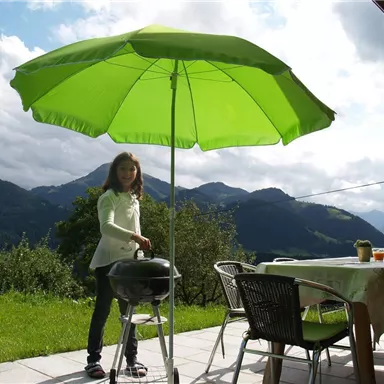 A girl is standing under a large green sun umbrella and grilling. In the background, mountains and a green meadow can be seen.