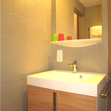 Ein modernes Badezimmer mit einem Waschbecken und Holzschrank. Auf dem Regal stehen bunte Becher und es gibt ein großes Spiegellicht.