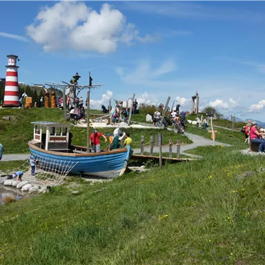 A picturesque viewpoint with a boat and a lighthouse in the background. Many people are enjoying nature and sitting on the grass.