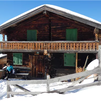 A traditional wooden mountain cabin with green shutters stands in the snow. The sky is clear and blue, and the surroundings are wintry.