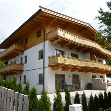 A modern building in alpine style with wooden cladding and balconies. It is situated in a green environment with trees and a garden.