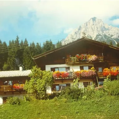 A traditional mountain hut with colorful flower balconies. In the background, high mountains rise beneath a blue sky.