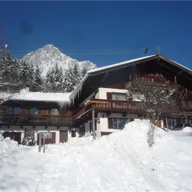 A traditional chalet in the snow, surrounded by trees. In the background, there is a snow-covered mountain and a radiant blue sky.