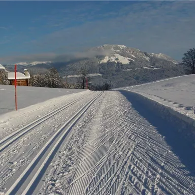 A snowy landscape with ski slopes and a clear blue sky. In the background, snow-covered mountains and some wooden houses can be seen.