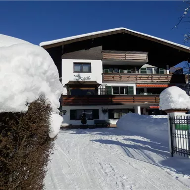 Ein gemütliches Holzhaus im Schnee mit Balkonen und einem strahlend blauen Himmel. Der Weg vor dem Haus führt durch eine verschneite Landschaft.