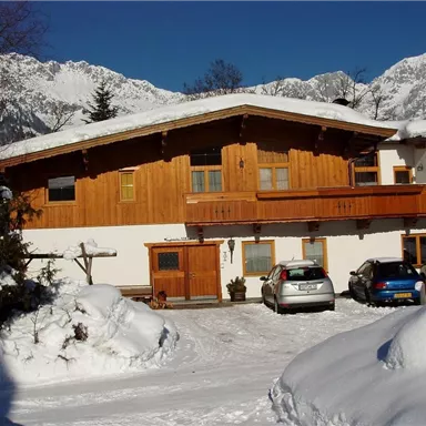A wooden house with a balcony, surrounded by snow and mountains. In the foreground, several cars are parked in a snowy parking lot.