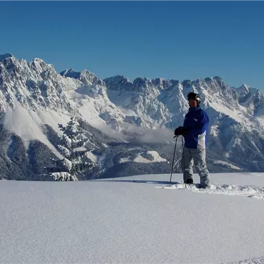 A man stands in a snow-covered landscape with high mountains in the background. The sky is clear and blue.