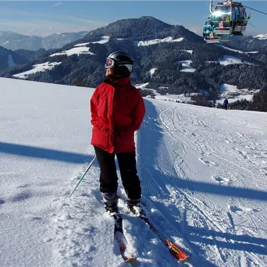 A person in red ski clothing stands on a snow-covered slope. In the background, mountains and a ski lift can be seen.