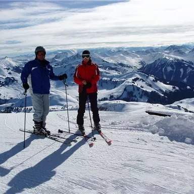 Two skiers are standing on a snowy slope. In the background, impressive mountains and a clear sky can be seen.