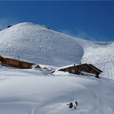 Two traditional huts are situated in a snow-covered landscape. In the background, a gentle hill stretches under a clear blue sky.