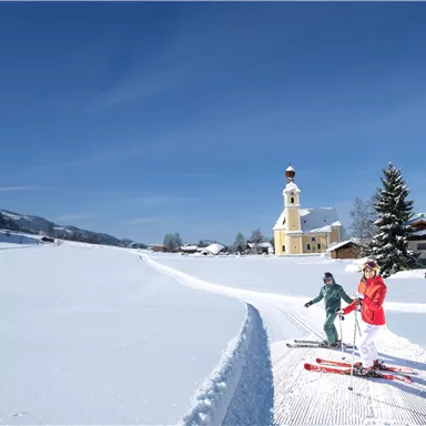 Two skiers are skiing through a snow-covered landscape. In the background, there is a picturesque church and a clear blue sky.