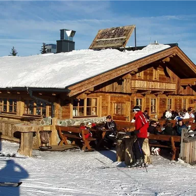 A cozy cabin in the snow with a snow-covered roof. Skiers and guests enjoy the sun on the terrace.