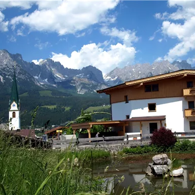 Eine malerische Landschaft mit Bergen im Hintergrund und einem ruhigen Teich im Vordergrund. Im Bild sind auch ein traditionelles Haus und ein Kirchturm zu sehen.