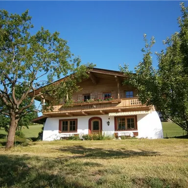 Ein traditionelles, zweigeschossiges Haus im bayerischen Stil mit Holzverandabalkon. Umgeben von grünen Wiesen und Bäumen unter klarem Himmel.