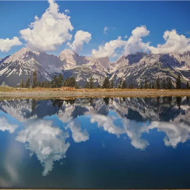 Eine beeindruckende Berglandschaft mit schneebedeckten Gipfeln und einem klaren, spiegelnden See. Der Himmel ist blau mit weißen Wolken.