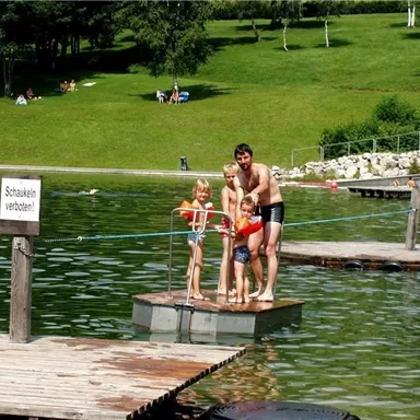 A man is standing with two children on a floating raft in the water. In the background, there are green meadows and other visitors at the lake.