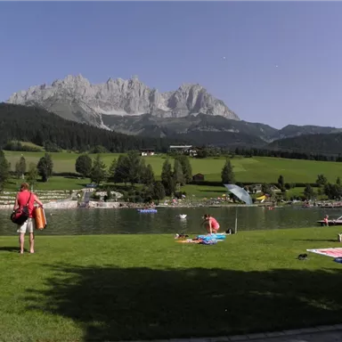 A picturesque lake surrounded by mountains and meadows. People enjoy the sun and relax on the shore.