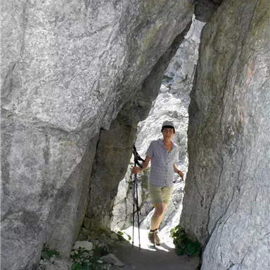 A person is stuck between large boulders and is holding a hiking stick. The path looks narrow and rough, surrounded by gray rock.