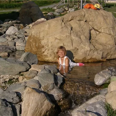 A child sits in the water between large stones and smiles. In the background, green meadows can be seen.