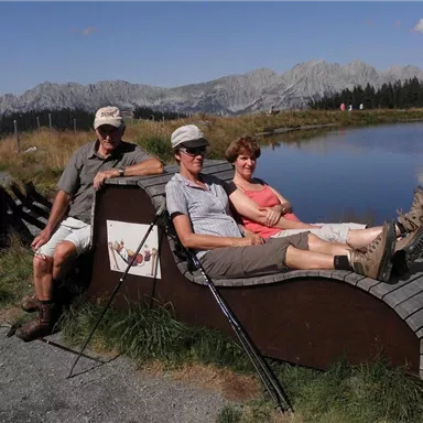 Three people are sitting relaxed on a wooden lounge chair by the shore of a lake. In the background, there are mountains and a clear blue sky.
