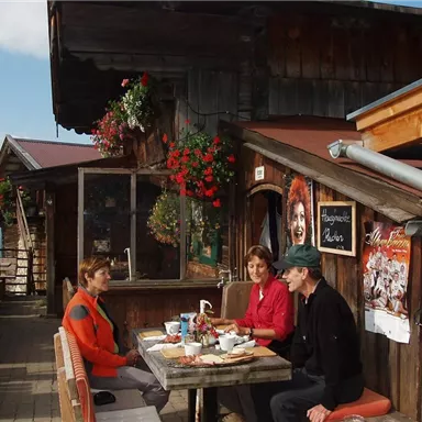 A cozy alpine cabin with blooming plants. Three people are sitting at a table and enjoying their breakfast outdoors.