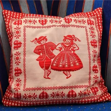Ein dekorisches Kissen mit roten, folkloristischen Motiven von tanzenden Paaren. Der Hintergrund des Kissens ist beige mit einem geometrischen Rahmen.