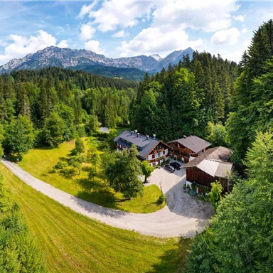 Eine malerische Landschaft mit Bergen und dichten Wäldern. Im Vordergrund stehen eine Ansammlung von Gebäuden und ein gewundener Weg.