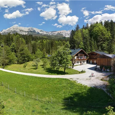 Eine malerische Panoramalandschaft mit einem Ferienhaus und umgebenden Wäldern. Im Hintergrund sind Berge und ein klarer blauer Himmel zu sehen.